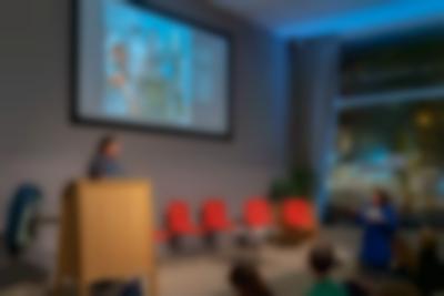 A person stands behind a podium on stage. They looks towards another person, who is off-stage to their right. The person off-stage looks as if they are speaking.