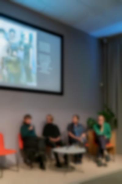 Four people sit on stage, around a low round table. A slide with a photograph and a short text is being projected behind them. The person to the left holds a microphone.