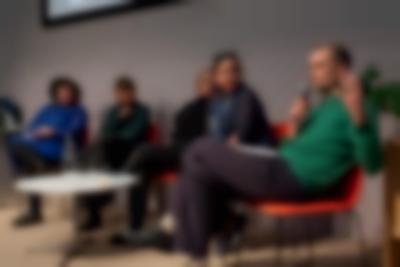Five people sit on stage, around a low round table. The person to the right holds a microphone and is gesturing with their left hand.