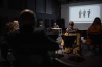 Several people, sitting on chairs, face the front of the room, where Megan is delivering a presentation. Behind her, a slide is being projected onto a screen.