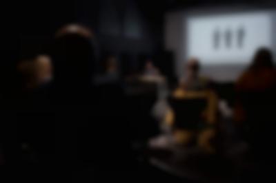 Several people, sitting on chairs, face the front of the room, where Megan is delivering a presentation. Behind her, a slide is being projected onto a screen.