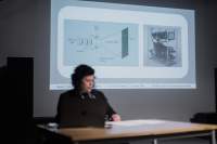 Megan sits behind a desk and a computer, delivering a presentation. Behind her, a slide is being projected.