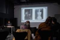 Several people, sitting on chairs, face the front of the room, where Megan is delivering a presentation. Behind her, a slide with two photographs is being projected onto a screen.