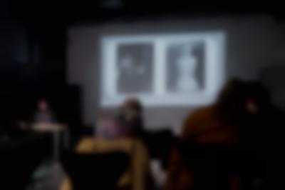 Several people, sitting on chairs, face the front of the room, where Megan is delivering a presentation. Behind her, a slide with two photographs is being projected onto a screen.