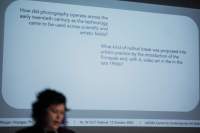 Megan's head appears in front of the powerpoint slide projection. The slide asks two questions about how the usage of photography and video throughout the twentieth century.