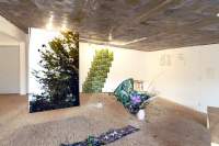 In an exhibition space, a large photographic print with green foliage is suspended from the ceiling. To the left, a photographic print has been placed on top of a plant in a vase. To the front of the print, a long line of flower prints that stretches across the floor is partially visible. On the walls, there are more prints with images and text.