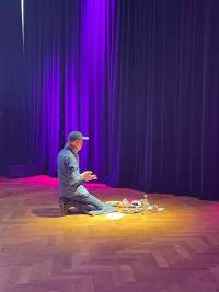 Sands Murray-Wassink sits on his heels in front of several art supplies. He faces the curtains, to the back of the space.