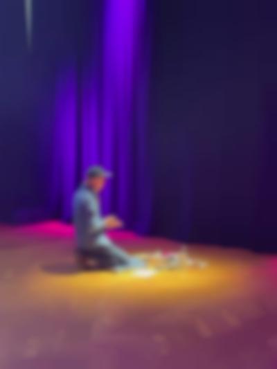 Sands Murray-Wassink sits on his heels in front of several art supplies. He faces the curtains, to the back of the space.