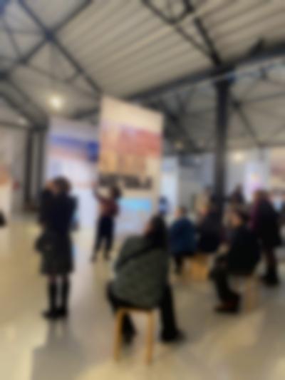 Lofty exhibition space with several large screens hanging from the ceiling. Samia Henni stands in front of one of the screens, pointing to the left. A group of people sitting in front, look in the direction Samia is pointing at.