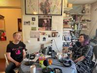 Sands Murray-Wassink and Megan Hoetger  are sitting across from each other at a table. They both have microphones in front of them. Behind them there is a bookshelf and a wall with posters, postcards and other decor.
