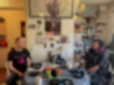 Sands Murray-Wassink and Megan Hoetger  are sitting across from each other at a table. They both have microphones in front of them. Behind them there is a bookshelf and a wall with posters, postcards and other decor.