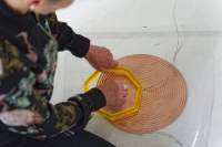 A coil spiral has been stuck to a white floor with a transparent plastic. There are two cables attached to it, one to the centre and the other to the edge. A person can partially be seen sitting next to it. With their left hand they are holding a small yellow heptagon structure against the spiral. With their right hand, they are touching the spiral through the middle of the structure.