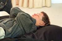 A person is lying supine on a large black cushion. They are wearing a grey sweater. Their hands are covering their ears and their eyes are closed.