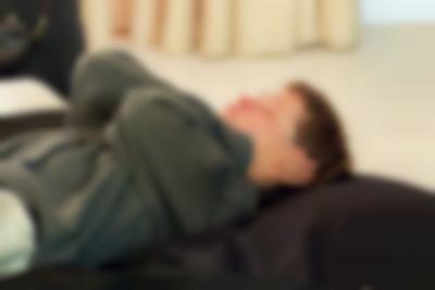 A person is lying supine on a large black cushion. They are wearing a grey sweater. Their hands are covering their ears and their eyes are closed.