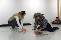 Two people seem to be playing with a sound installation consisting of two coil spirals stuck to the floor. There are cables attached to both spirals. A booklet is open next to one of them. In the background, a person is laying on the floor. They have their elbows on a pillow and are reading a book or a magazine.