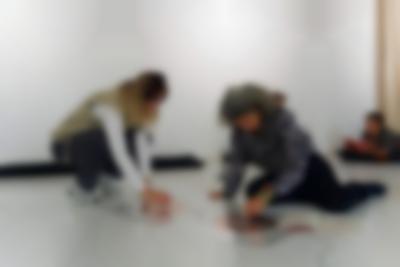 Two people seem to be playing with a sound installation consisting of two coil spirals stuck to the floor. There are cables attached to both spirals. A booklet is open next to one of them. In the background, a person is laying on the floor. They have their elbows on a pillow and are reading a book or a magazine.