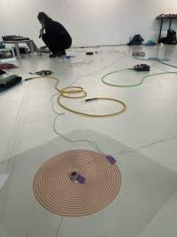 In the backgroud, a person dressed in black is squatting in front of a sound deck and a computer, which they are using. They are holding a microphone to their mouth. At the front, a red coil spiral is stuck to the floor. A thin cable is attached to its center with purple table. Throughout the rest of the space, other sound equipment and colourful cables lay on the flor.