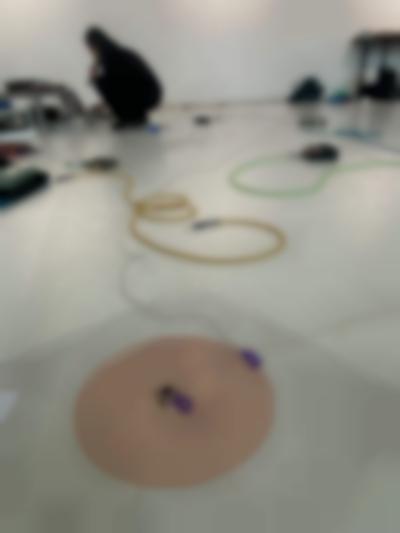 In the backgroud, a person dressed in black is squatting in front of a sound deck and a computer, which they are using. They are holding a microphone to their mouth. At the front, a red coil spiral is stuck to the floor. A thin cable is attached to its center with purple table. Throughout the rest of the space, other sound equipment and colourful cables lay on the flor.