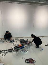 Decks and other sound equipment have been set in two low tables and on the ground. A person can be seen squatting next to the larger structure, using a laptop. Someone else is sat by the wall, with a laptop to the left and a deck to the right, which they are adjusting.