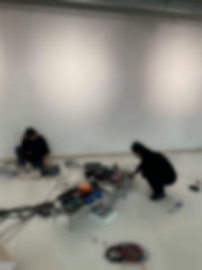 Decks and other sound equipment have been set in two low tables and on the ground. A person can be seen squatting next to the larger structure, using a laptop. Someone else is sat by the wall, with a laptop to the left and a deck to the right, which they are adjusting.