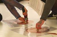 Close-up of two coil spirals, stuck to the white ground with a transparent plastic. There are wires coming from each of these spirals, and another wire connecting them both. The arms and a foot of two people can be seen. One person holds a microphone to a magnet, that they have set on one of the spirals. The other person touches the centre of the other spiral.