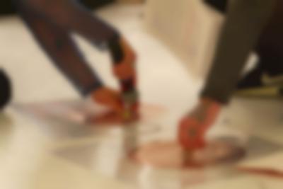 Close-up of two coil spirals, stuck to the white ground with a transparent plastic. There are wires coming from each of these spirals, and another wire connecting them both. The arms and a foot of two people can be seen. One person holds a microphone to a magnet, that they have set on one of the spirals. The other person touches the centre of the other spiral.