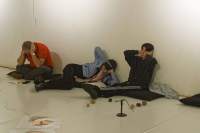 Three people are sitting or laying on black cushions on the floor, next to a white wall. They are sitting in different positions, but are all holding their hands to their ears.