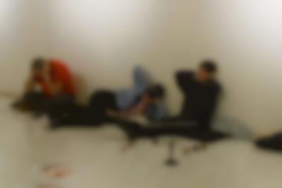 Three people are sitting or laying on black cushions on the floor, next to a white wall. They are sitting in different positions, but are all holding their hands to their ears.