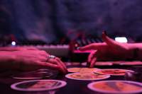 Two hands, belonging to two people sitting across from each other, are touching the same tarot card. Around it, there are other cards.