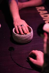A hand hovers over a small basket, possibly made from cotton or similar material. The basket is set on a dark table. The hand of someone sitting across rests on the table.