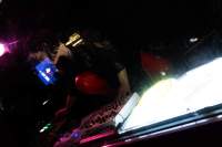 A person stands behind a DJ booth. They are adjusting one of the decks. Next to the decks there is a light box, which emits a bright white light.