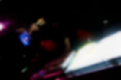 A person stands behind a DJ booth. They are adjusting one of the decks. Next to the decks there is a light box, which emits a bright white light.