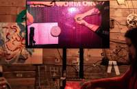 A screen on a stand shows a table filmed from above. The arms of two people, sitting across from each other, rest on the table. There is also a sheet of paper with handwritten notes and a stack of round tarot cards. The screen is set in a wood-panelled room with some wall decoration.