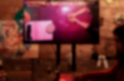 A screen on a stand shows a table filmed from above. The arms of two people, sitting across from each other, rest on the table. There is also a sheet of paper with handwritten notes and a stack of round tarot cards. The screen is set in a wood-panelled room with some wall decoration.