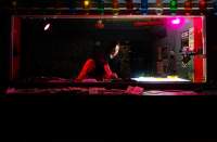 A person stands in a dark DJ booth, adjusting the knobs on a DJ deck. To their right, a bright light shines upwards from a light box set next to the deck.