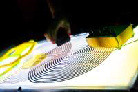 On top of a light box surface, there are two coil spirals and a small sound deck with knobs. A hand can be seen holding an object, which they've put on top of one of the spirals.