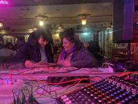 Next to a sound deck, two people are looking through a newspaper and smiling. They are in an indoor space after dark, possibly a bar or a cafe.