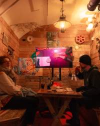 Two people sit across from each other. In between them there is a table with drinks and paper materials. One of the people is taking a sip from a beer. At the back, there is a screen on a stand, showing a table filmed from above. In this image, someone rests their arms on the table, with their palms facing up. Someone is sitting across from them. There is a sheet of paper and a stack of round tarot cards.