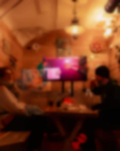 Two people sit across from each other. In between them there is a table with drinks and paper materials. One of the people is taking a sip from a beer. At the back, there is a screen on a stand, showing a table filmed from above. In this image, someone rests their arms on the table, with their palms facing up. Someone is sitting across from them. There is a sheet of paper and a stack of round tarot cards.