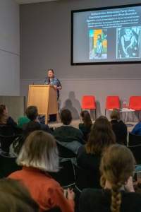 Nearly full lecture room. On stage, a person stands behind a podium. There is slide being projected behind them. It has a text, a painting and a photograph in black-and-white.