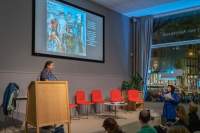 A person stands behind a podium on stage. They looks towards another person, who is off-stage to their right. The person off-stage looks as if they are speaking.