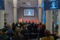 Nearly full lecture room. On stage, a person stands behind a podium. There is an image being projected on stage and in another screen, which is closer to the middle of the room. The image shows a feminine-presenting person with short hair in black-and-white.