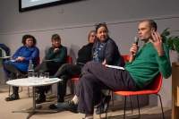 Five people sit on stage, around a low round table. The person to the right holds a microphone and is gesturing with their left hand.