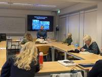 In a university room, four people sit around tables set up in a U shape. At the front, a person is standing up or slightly resting against a table. A slide with two black and white photographs is being projected behind them.