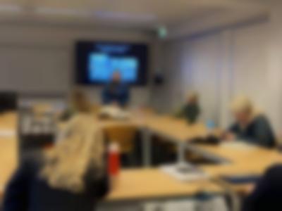 In a university room, four people sit around tables set up in a U shape. At the front, a person is standing up or slightly resting against a table. A slide with two black and white photographs is being projected behind them.