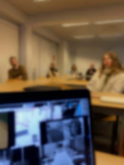 A laptop screen is partially visible, with several  photo files open on the desktop. Behind it, five people sit around tables in a university room.