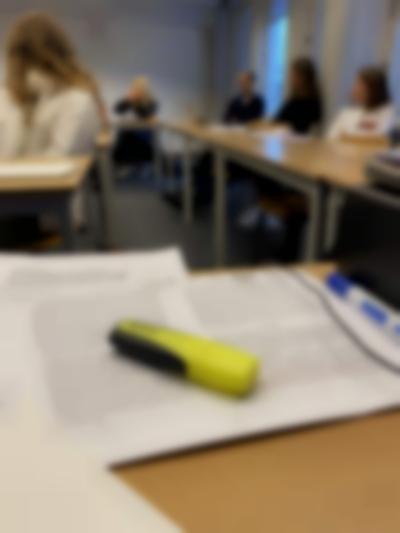 Two text print outs have been set on a table. A thick yellow highlighter lays on top. In the background, five other people sit around other tables, looking in the same direction.