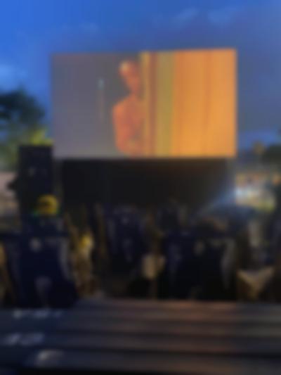 Several deck chairs face a large outdoor screen, onto which a film is being projected. On screen, a feminine-presenting person rests their face on a door frame.